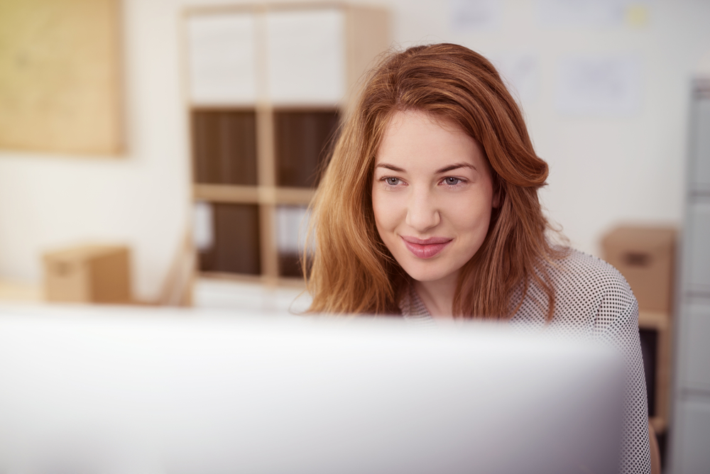 Wanita muda yang menarik bekerja di komputer desktop sambil tersenyum saat dia mencondongkan tubuh ke depan membaca teks di layar, melihat ke atas monitor